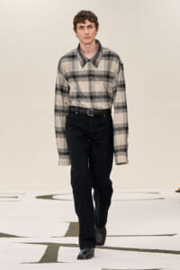 Male model walking a runway in a beige-gray plaid button-down shirt, black belt, and black jeans with black boots?
