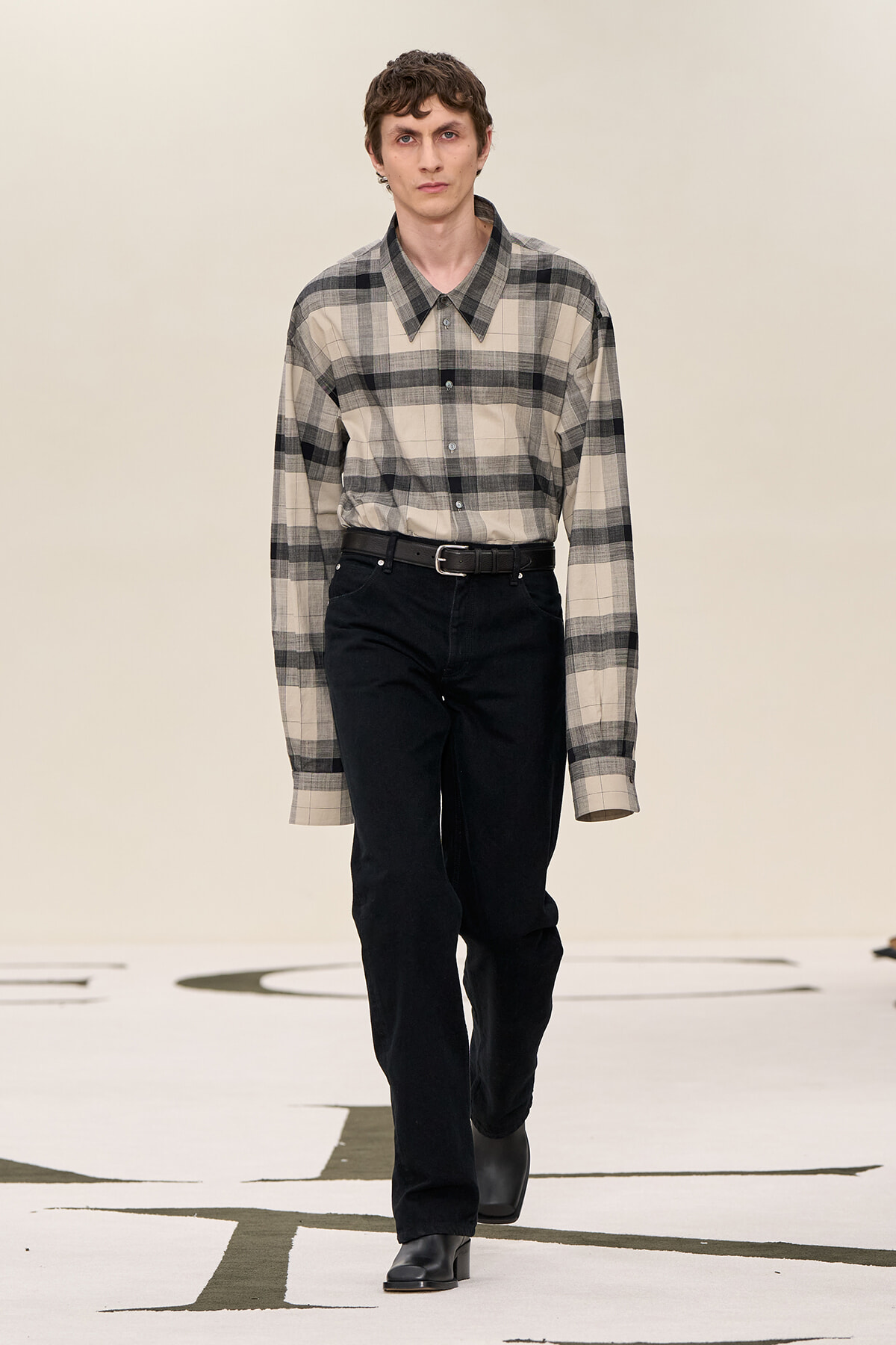 Male model walking a runway in a beige-gray plaid button-down shirt, black belt, and black jeans with black boots?