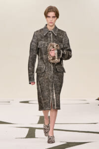 Model walking a runway in a gray textured tweed coat and matching midi skirt, holding a brown patterned bag.