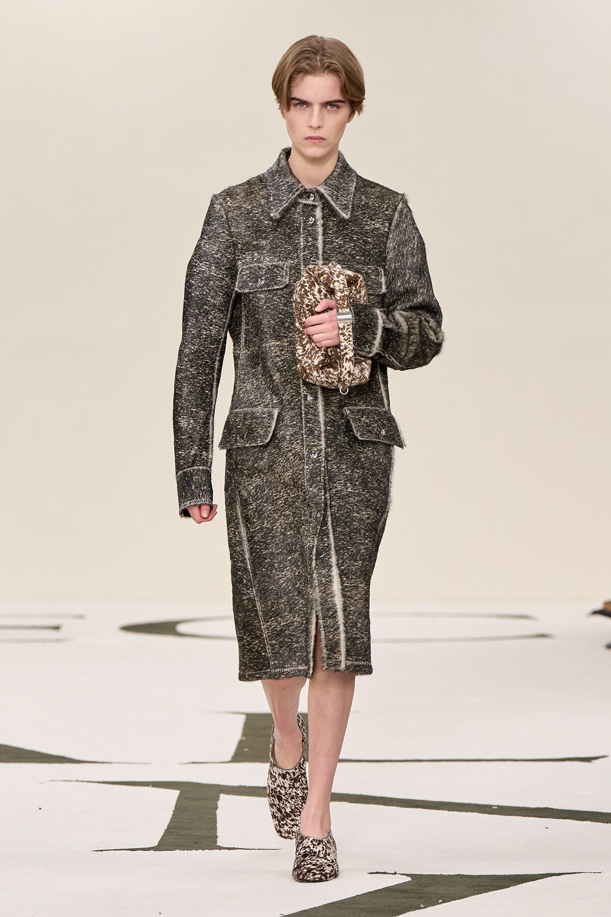 Model walking a runway in a gray textured tweed coat and matching midi skirt, holding a brown patterned bag.