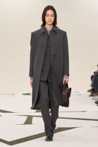 Model walking a runway in a dark gray oversized coat over a matching suit, carrying a brown structured handbag.
