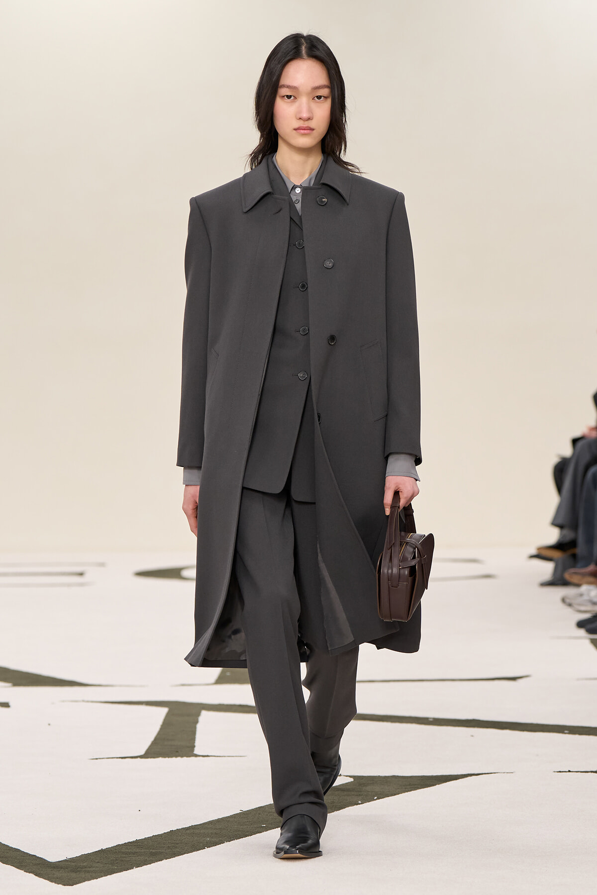 Model walking a runway in a dark gray oversized coat over a matching suit, carrying a brown structured handbag.