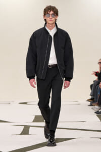 Male model walking a fashion runway in a black bomber jacket, white shirt, black trousers, and shiny black shoes, wearing sunglasses.