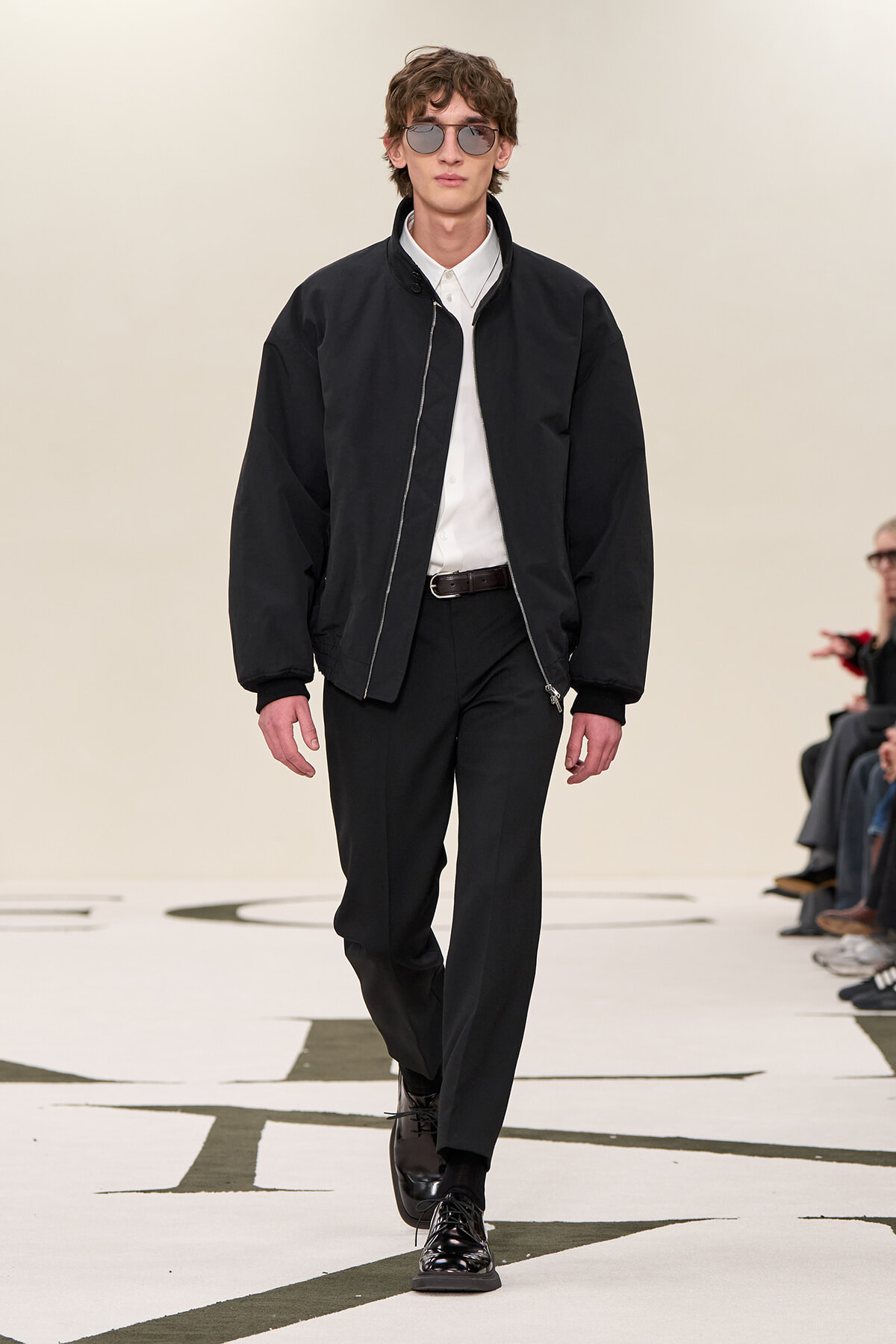 Male model walking a fashion runway in a black bomber jacket, white shirt, black trousers, and shiny black shoes, wearing sunglasses.