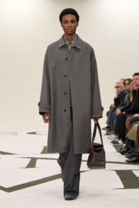 Male model on a fashion runway wearing a gray overcoat and matching trousers, carrying a brown leather bag as spectators watch from the side.
