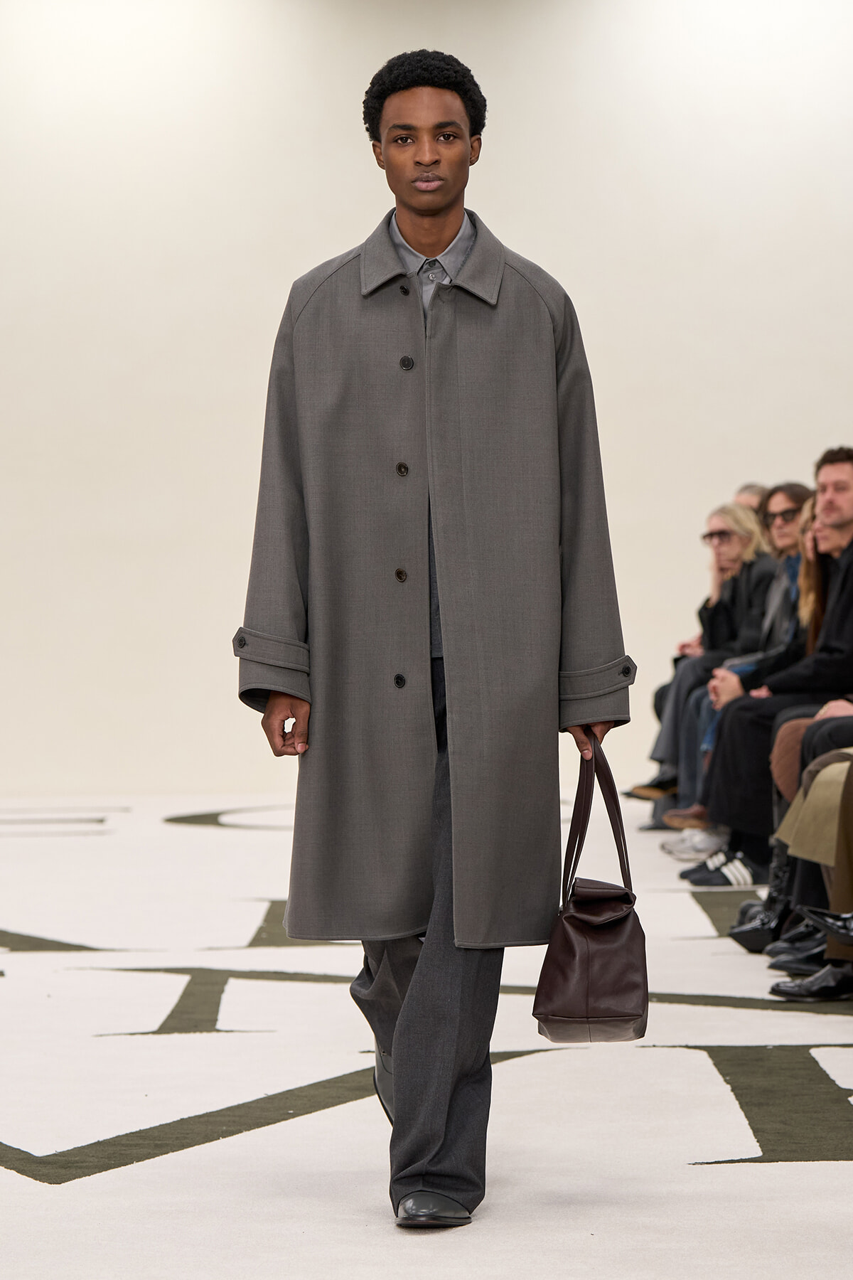 Male model on a fashion runway wearing a gray overcoat and matching trousers, carrying a brown leather bag as spectators watch from the side.