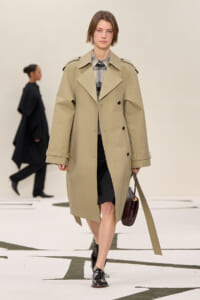 Model walking a runway in a beige trench coat over a plaid shirt and black skirt, carrying a burgundy handbag.
