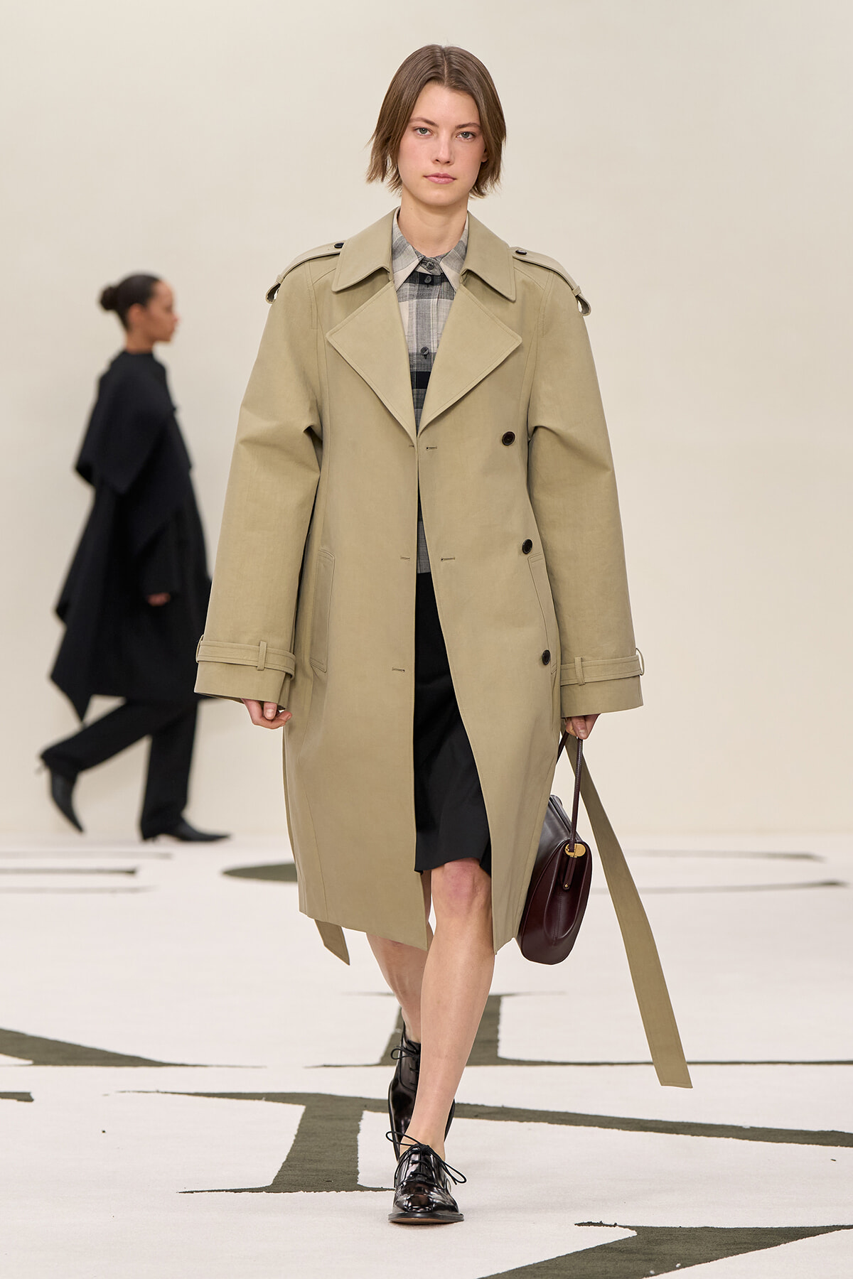 Model walking a runway in a beige trench coat over a plaid shirt and black skirt, carrying a burgundy handbag.