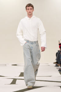 Male fashion model walking a runway in an oversized white hoodie and light-blue wide-leg jeans, audience seated along the sides.