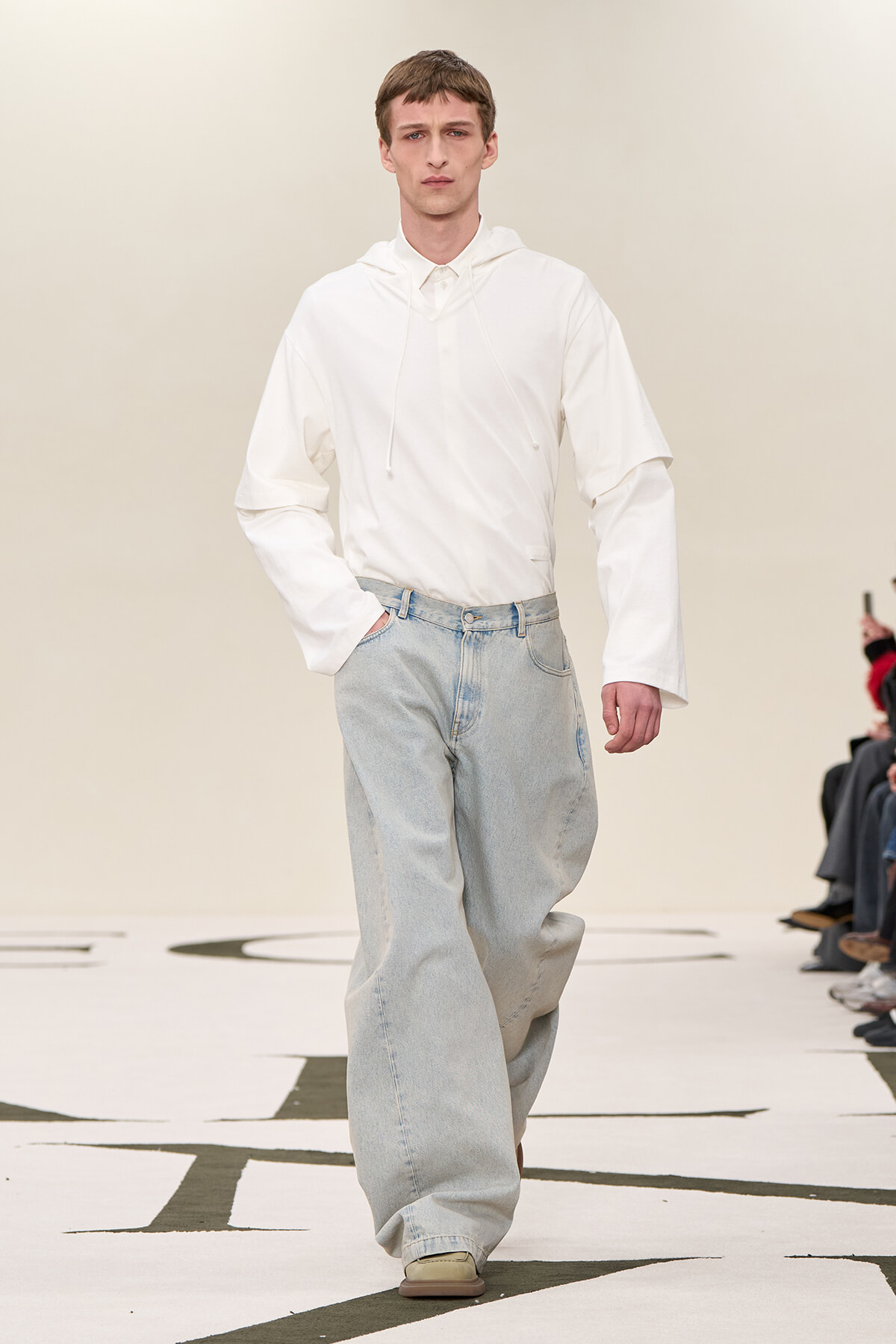 Male fashion model walking a runway in an oversized white hoodie and light-blue wide-leg jeans, audience seated along the sides.