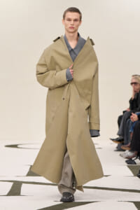Runway model wearing a long beige trench coat over a gray shirt, adjusting the coat as he walks.