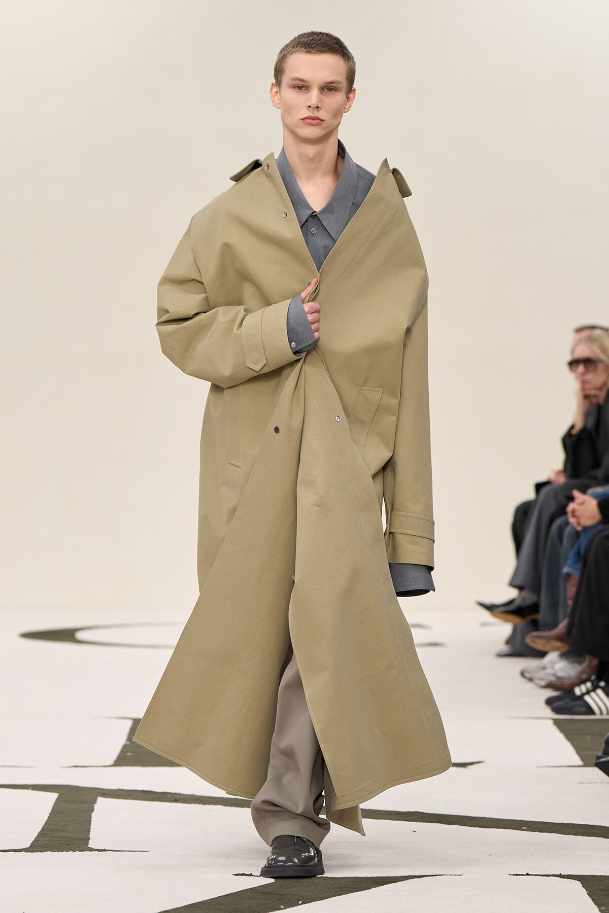 Runway model wearing a long beige trench coat over a gray shirt, adjusting the coat as he walks.