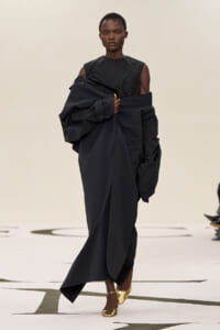 Model on a runway wearing an oversized black coat draped over a black sleeveless top and gold flats, creating a sculptural silhouette.