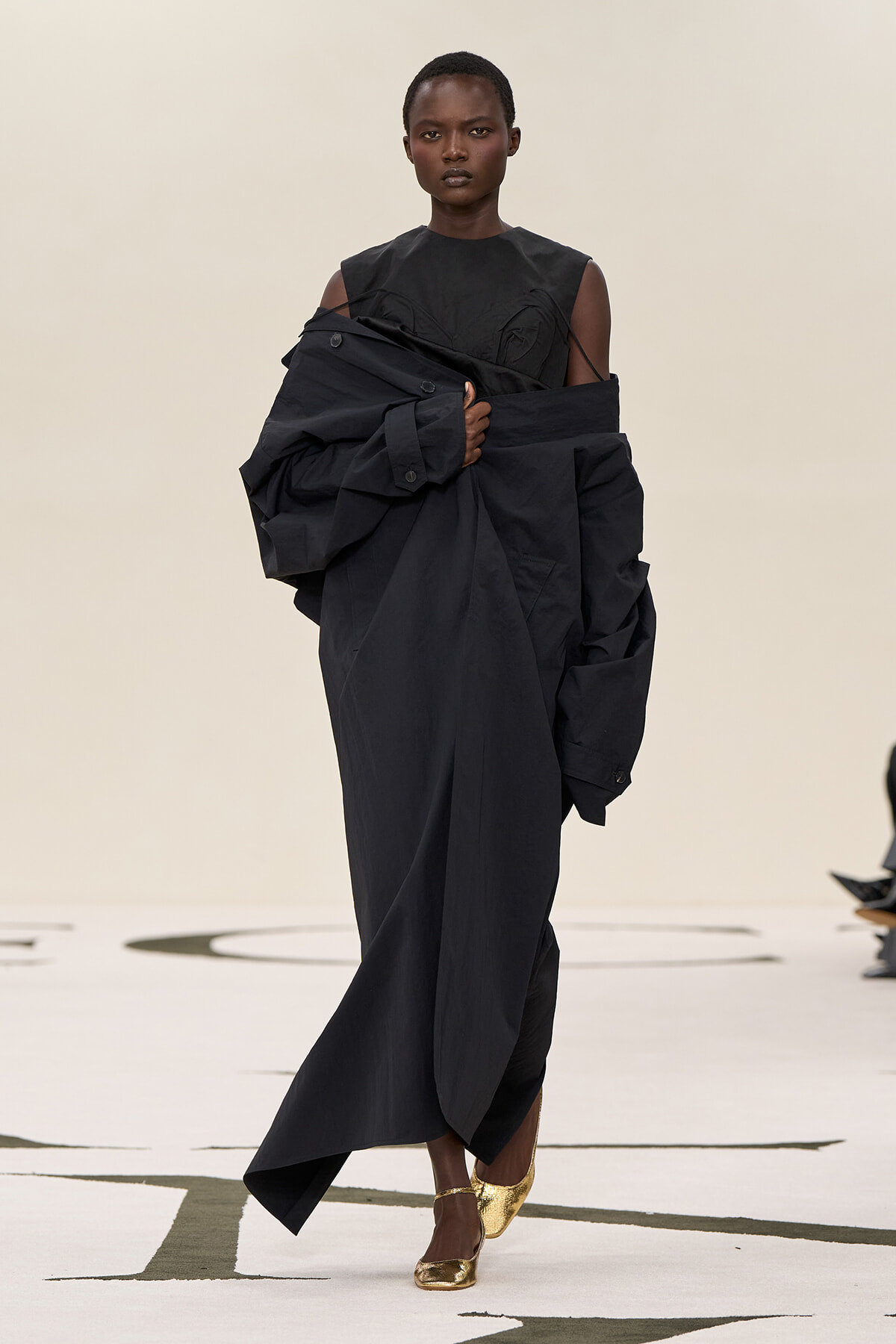 Model on a runway wearing an oversized black coat draped over a black sleeveless top and gold flats, creating a sculptural silhouette.
