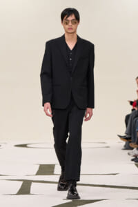 Male model walking a runway in a black tailored suit with a vest, sunglasses, and shiny dress shoes.