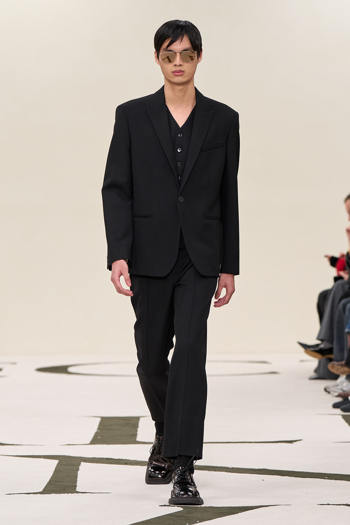 Male model walking a runway in a black tailored suit with a vest, sunglasses, and shiny dress shoes.