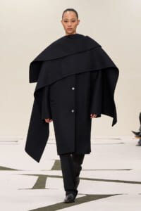 Model on runway wearing an oversized black cape coat with a wide, layered collar.