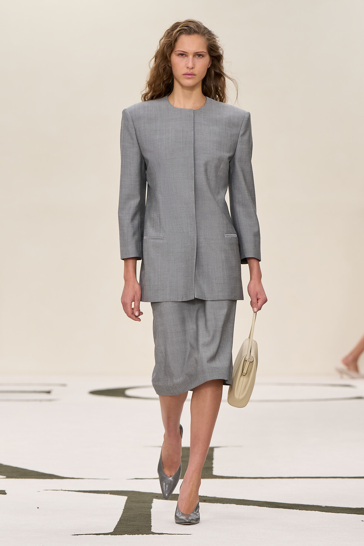 Model walks the runway in a tailored light gray blazer and matching knee-length skirt, holding a beige handbag.