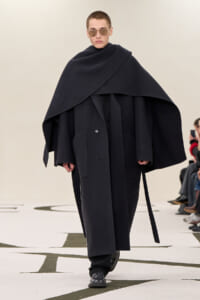 Model on runway wearing an oversized black cape over a long coat with wide collar and sunglasses. Conveys a minimalist, dramatic fashion look on a catwalk.