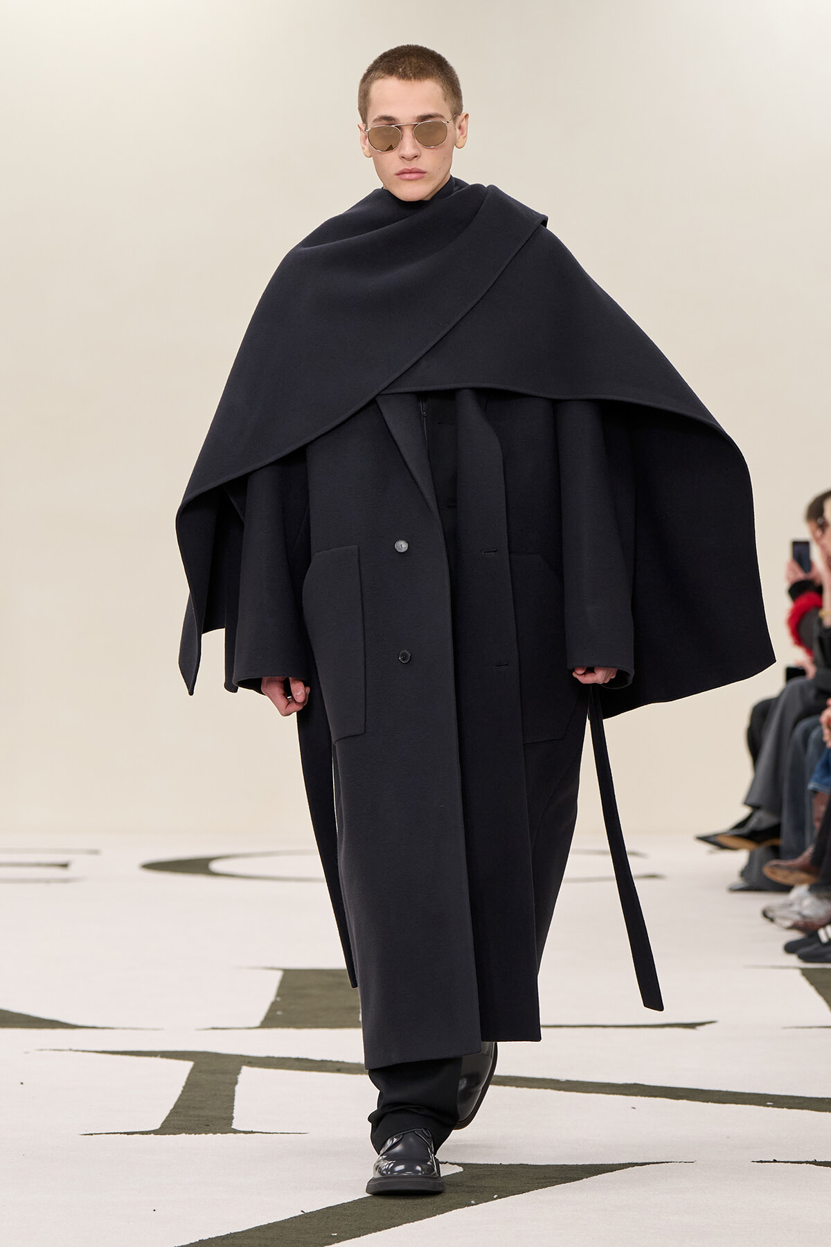 Model on runway wearing an oversized black cape over a long coat with wide collar and sunglasses. Conveys a minimalist, dramatic fashion look on a catwalk.