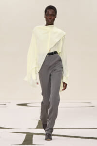 Model walking a runway in a pale yellow oversized blouse and gray tailored pants with a black belt.