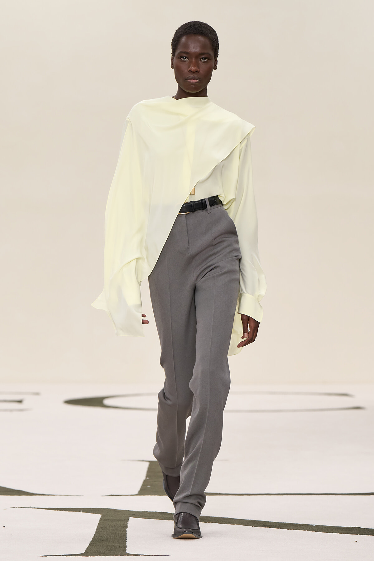 Model walking a runway in a pale yellow oversized blouse and gray tailored pants with a black belt.