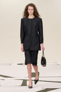 Model walking the runway in a black pinstripe suit with a structured blazer and knee-length skirt, carrying a black handbag.