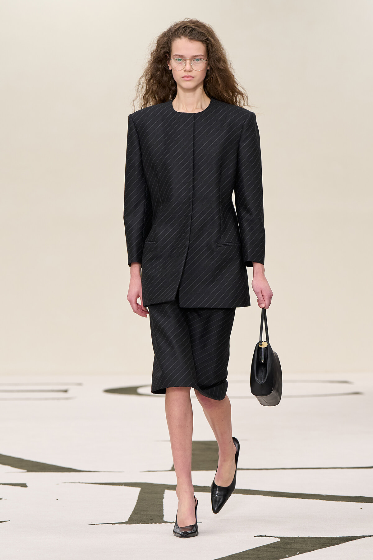 Model walking the runway in a black pinstripe suit with a structured blazer and knee-length skirt, carrying a black handbag.