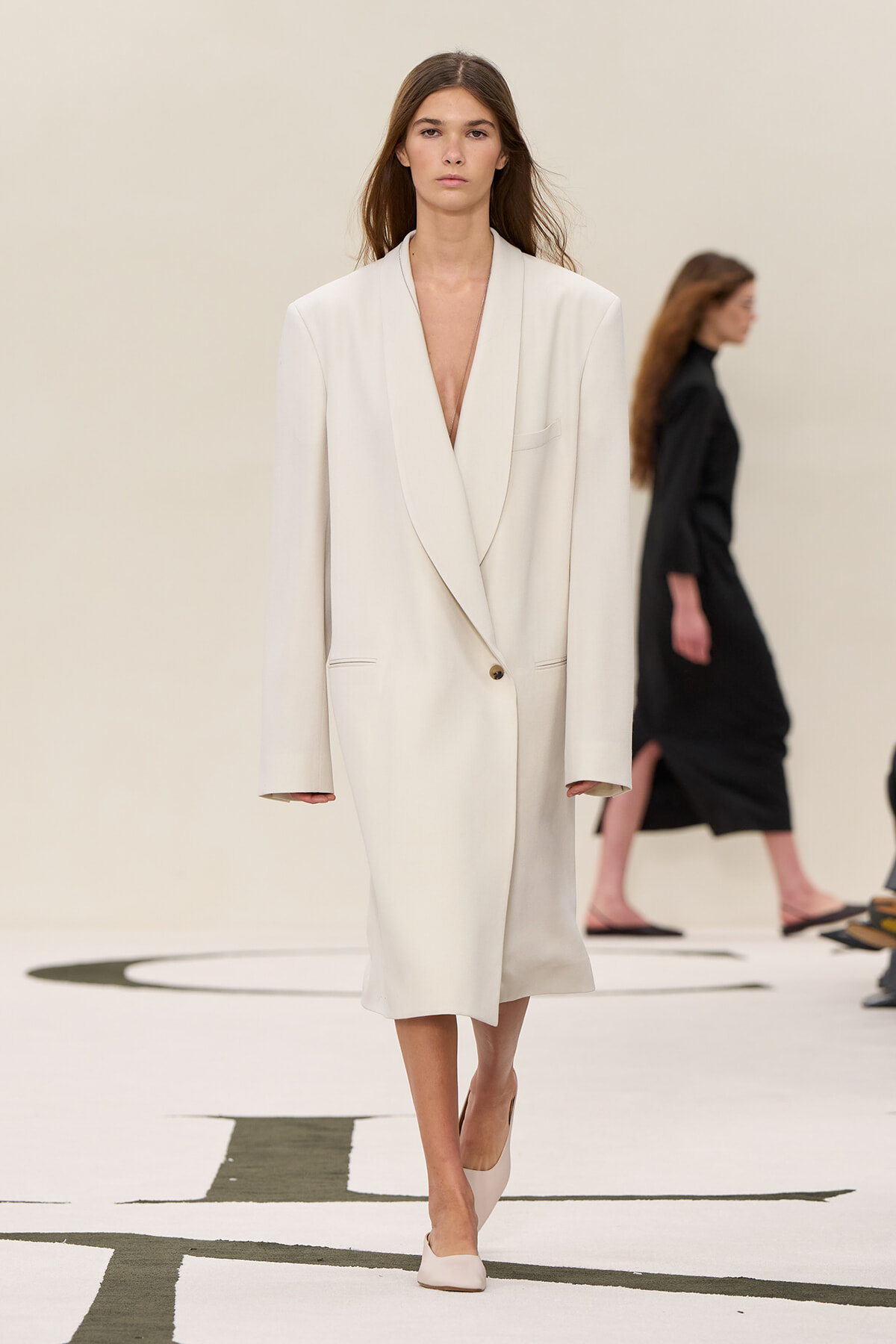 Model walks the runway in a cream oversized blazer dress with a plunging neckline and a single button fastening.