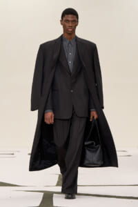Male model walking a runway in a dark pinstripe suit and long overcoat, carrying a black bag.