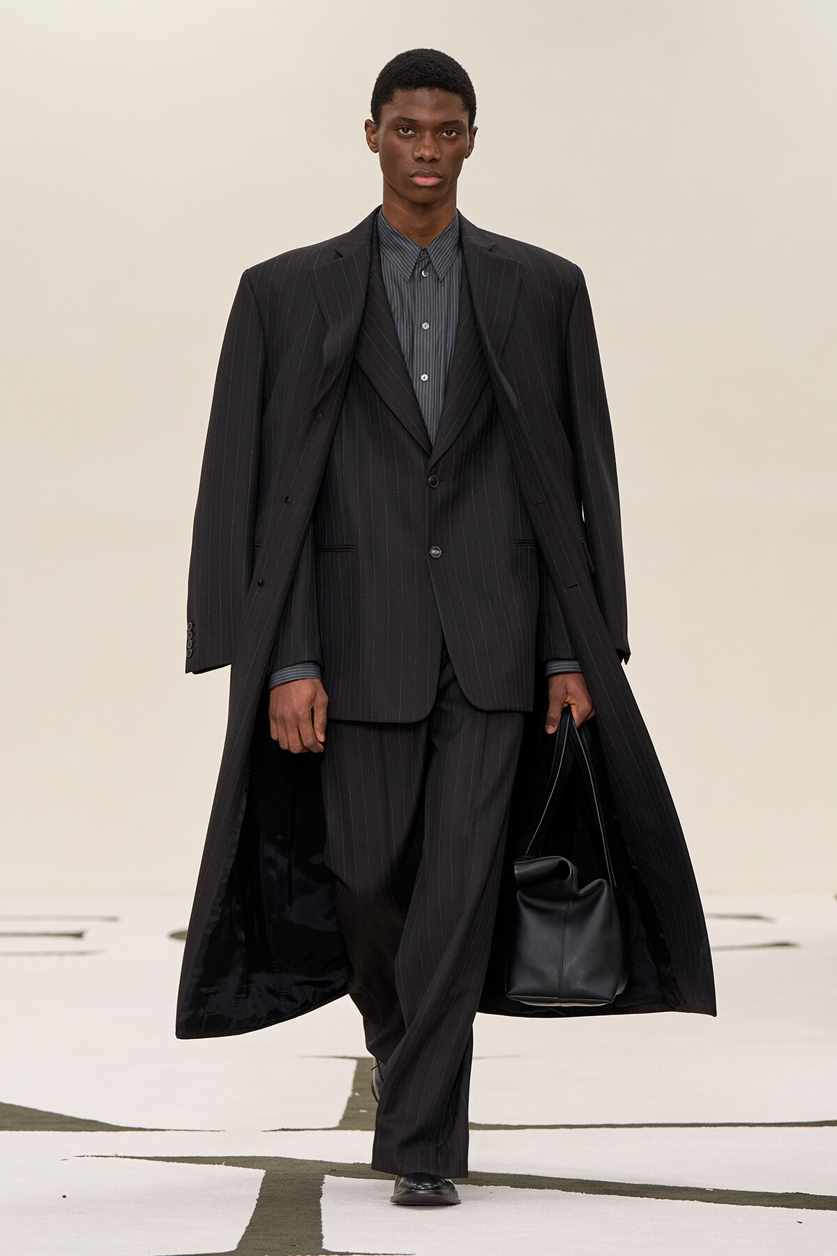 Male model walking a runway in a dark pinstripe suit and long overcoat, carrying a black bag.