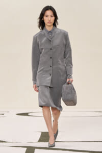 Model wearing a grey satin blazer and matching knee-length skirt, carrying a textured grey handbag while walking on a runway.