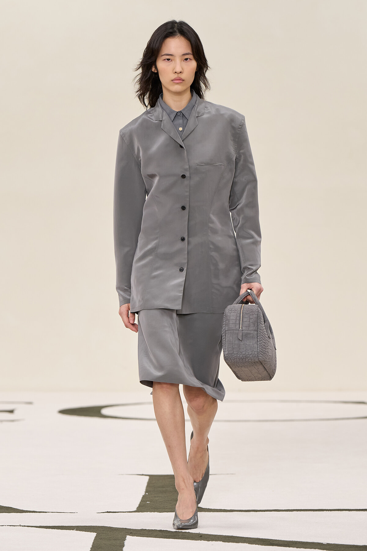Model wearing a grey satin blazer and matching knee-length skirt, carrying a textured grey handbag while walking on a runway.