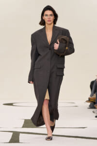 Model walks the runway in an oversized dark gray suit, carrying a brown quilted bag under her arm with glasses on.
