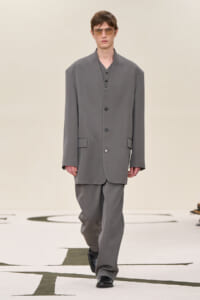 Male model wearing an oversized gray suit with a long blazer and wide trousers walking down a fashion runway, sunglasses on.