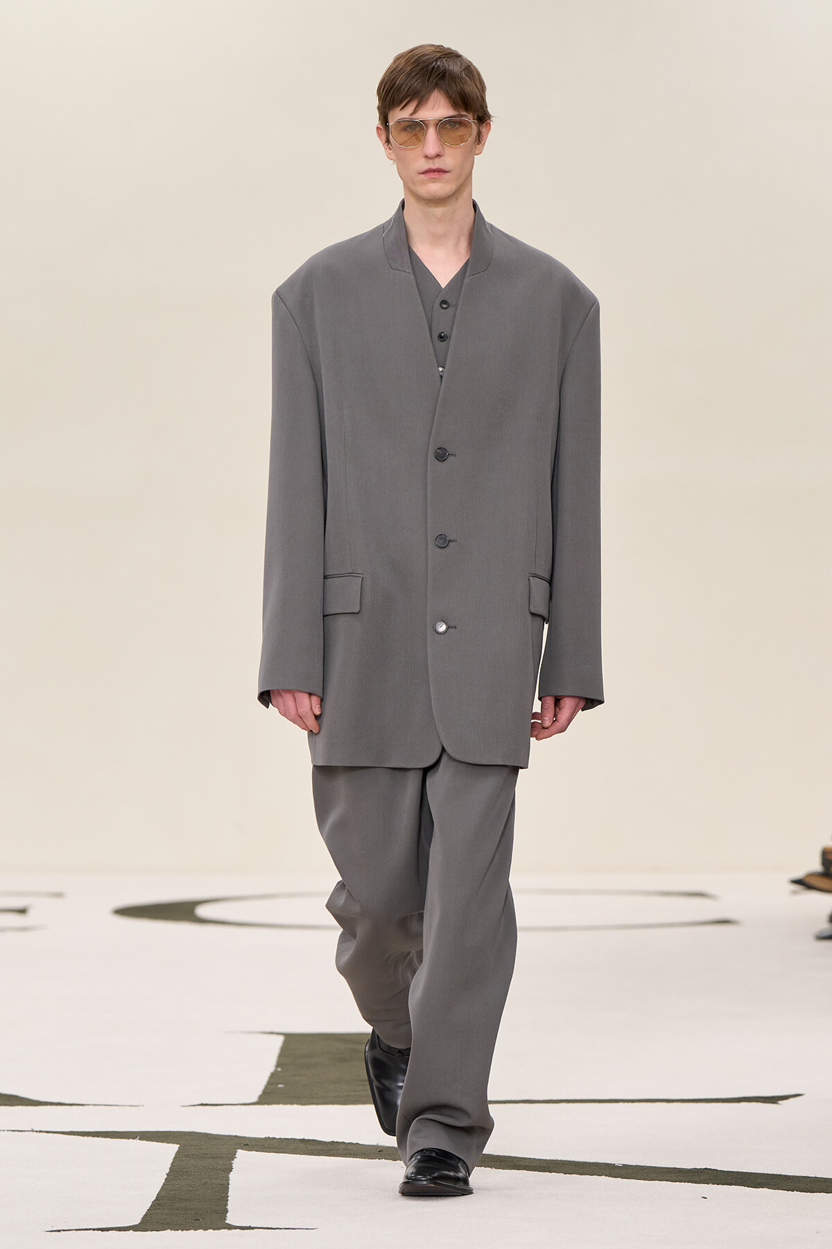 Male model wearing an oversized gray suit with a long blazer and wide trousers walking down a fashion runway, sunglasses on.