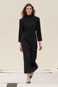 Model wearing a black high-neck midi dress with long sleeves, walking on a runway, wearing light glasses.