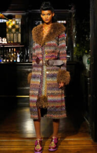 Model wearing a long multicolor zigzag coat with brown fur collar and cuffs, cinched by a wide belt, indoors at a bar.