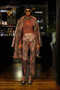 Model wearing a shimmering metallic red-gold suit with a matching cape, green beaded necklace, red pendant, and oversized cat-eye sunglasses in a bar setting.