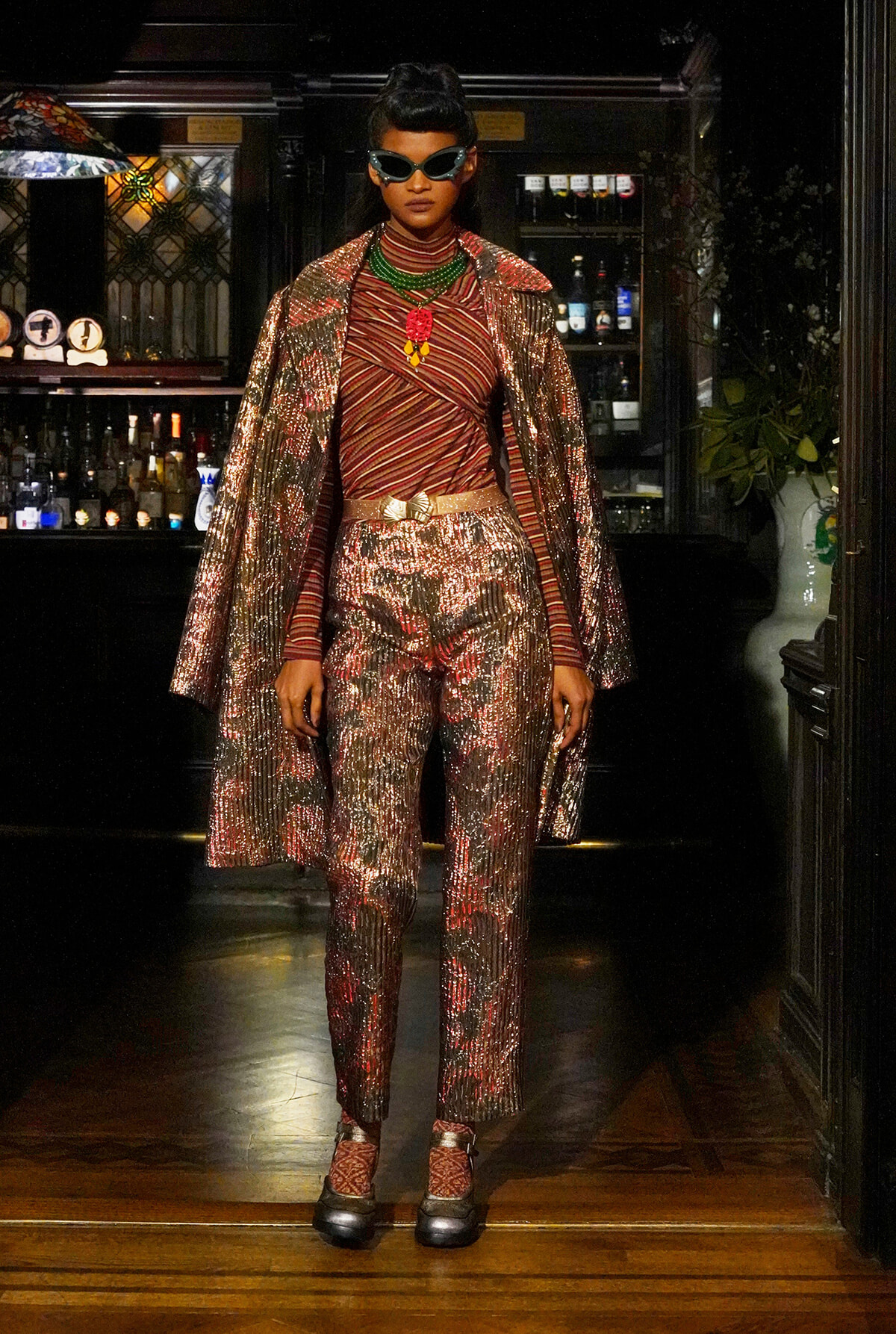 Model wearing a shimmering metallic red-gold suit with a matching cape, green beaded necklace, red pendant, and oversized cat-eye sunglasses in a bar setting.