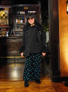 Woman in a black fur-trimmed coat and teal patterned skirt posing inside a bar.