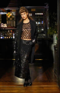 Woman in a black sequined outfit and coat stands in a dim bar, wearing a sheer dotted top and a wide belt with a circular buckle.