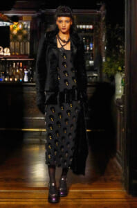 Woman in a black velvet coat over a black dress with gold circular embellishments, standing in a dim bar backdrop.