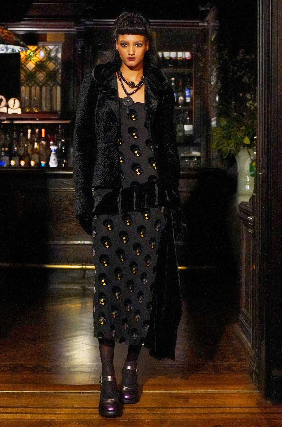Woman in a black velvet coat over a black dress with gold circular embellishments, standing in a dim bar backdrop.