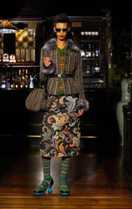 Model in a textured coat with fur cuffs and a colorful patterned skirt, standing in a bar with a matching tweed bag and sunglasses.
