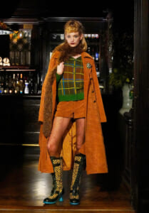 Model posing in a dim bar wearing an orange corduroy coat and matching shorts over a green plaid cardigan with a fuzzy scarf and leopard-print boots.