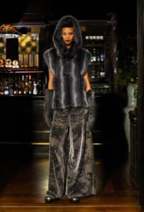 Full-length portrait of a model in a gray fur hooded vest with long fur cuffs and damask-patterned wide-leg pants, standing in a dim bar setting.