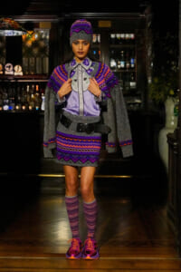 Model wearing a purple-gray knit cape, hat, and matching skirt with striped socks on a runway in a dim bar interior.