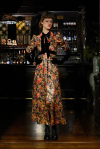 Woman in a long, floral vintage dress with black gloves and a net veil, standing in a dimly lit bar.