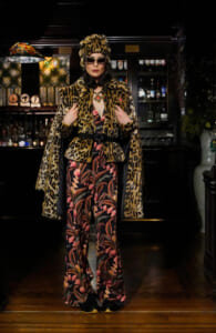 Person wearing a leopard print fur coat, matching hat and sunglasses posing in a bar setting with bottles in the background.